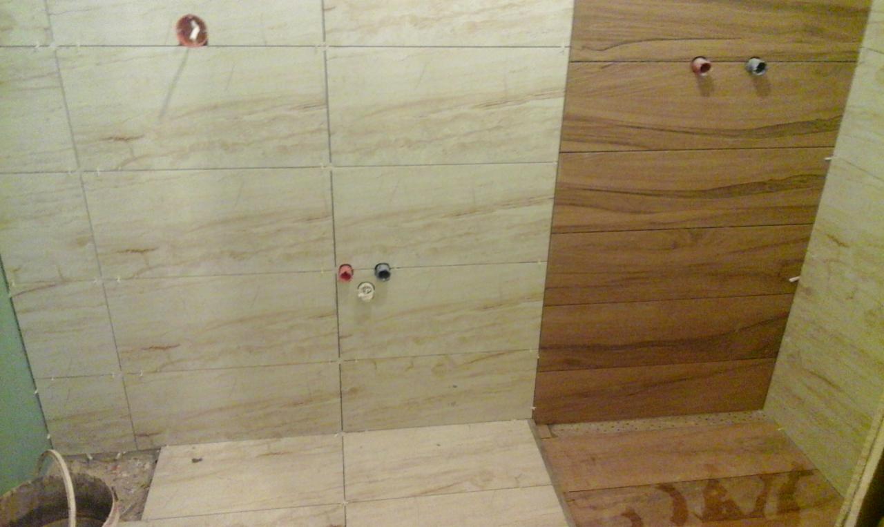 Ściana w trakcie układania płytek imitujących drewno i beżowych płytek z marmurkowym wzorem, widoczne wystające rury instalacyjne i plastikowe krzyżyki dystansowe.