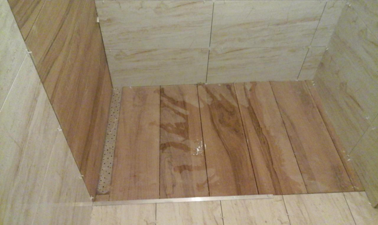 Wnętrze kabiny prysznicowej w trakcie wykańczania, z drewnopodobnymi płytkami na podłodze i beżowymi płytkami imitującymi marmur na ścianach, widoczne listwy wykończeniowe i elementy mocujące płytki.