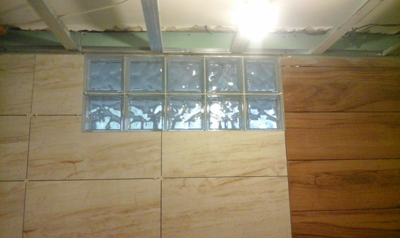 Fragment ściany wykończonej beżowymi płytkami ceramicznymi, z wstawką z dziewięciu szklanych luksferów, widoczny sufit w trakcie montażu