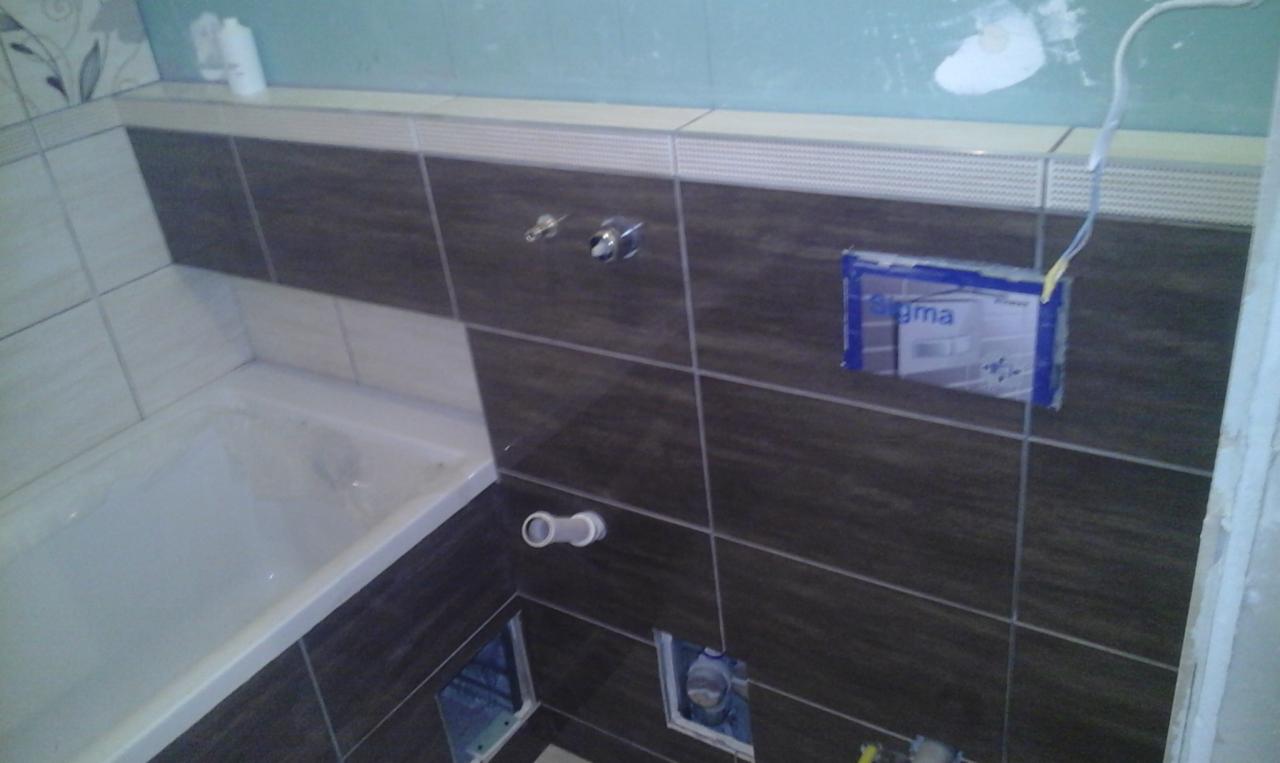 Fragment łazienki w trakcie wykańczania: brązowe i białe płytki częściowo ułożone wokół wanny, widoczne otwory instalacyjne i puszka podtynkową zabezpieczona taśmą.