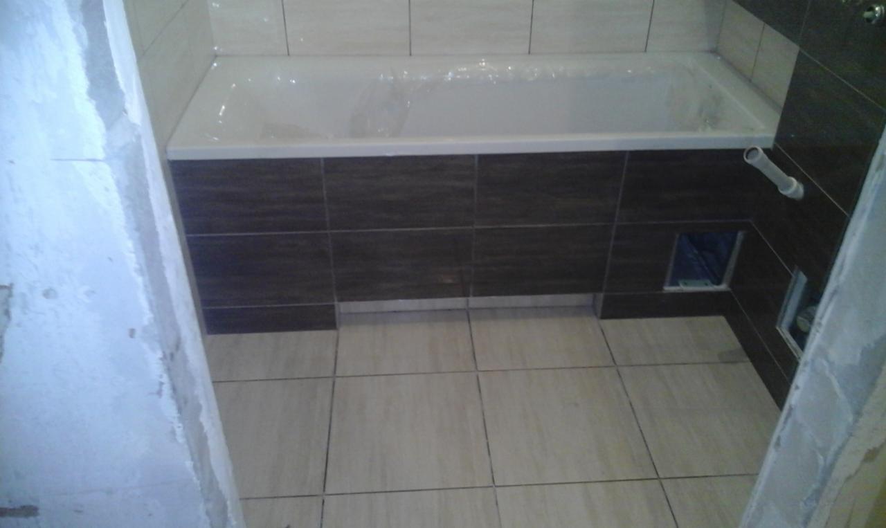 Biała wanna z brązową obudową z płytek, jasne płytki podłogowe i fragment ściany w trakcie wykończenia, widoczne otwory rewizyjne w obudowie wanny.