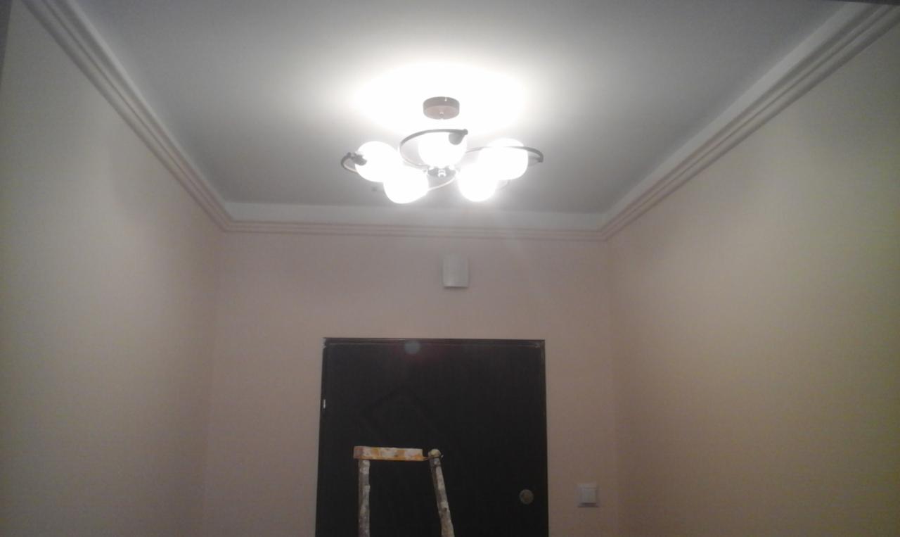 Pomieszczenie po remoncie: świeżo pomalowane ściany w odcieniach beżu, ozdobny gzyms pod sufitem, nowoczesna lampa sufitowa z kulistymi kloszami, drabina malarska oparta o ciemne drzwi.