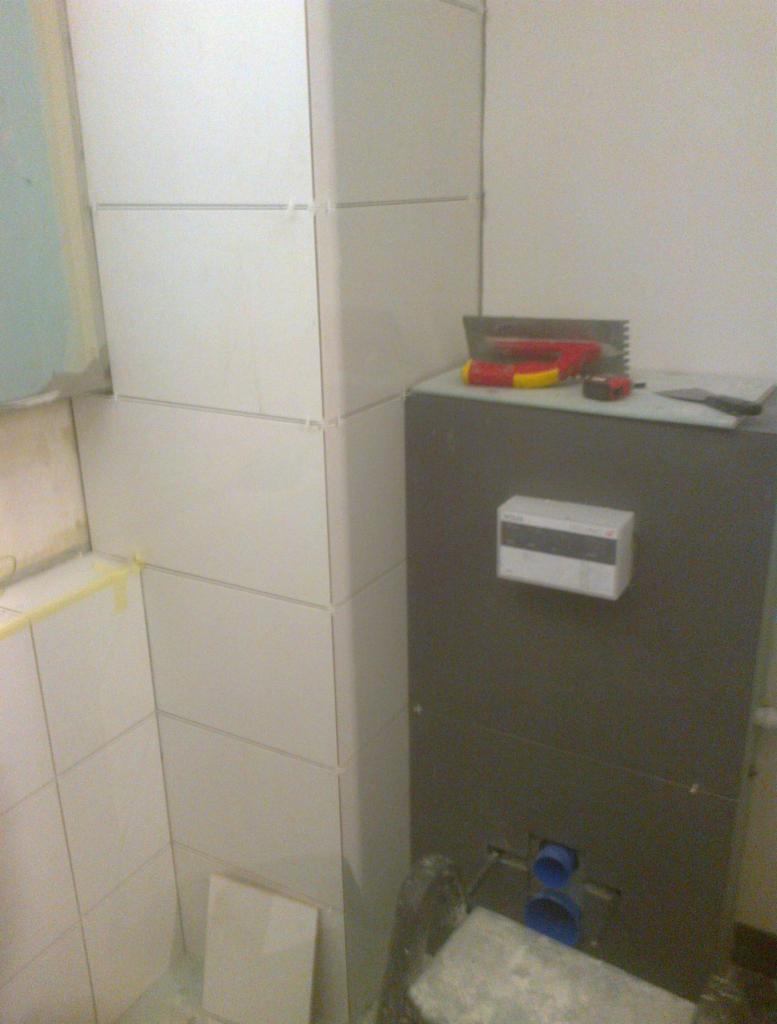 Białe płytki ceramiczne ułożone na rogu w łazience, widoczne narzędzia glazurnicze na szarej obudowie stelaża podtynkowego WC.