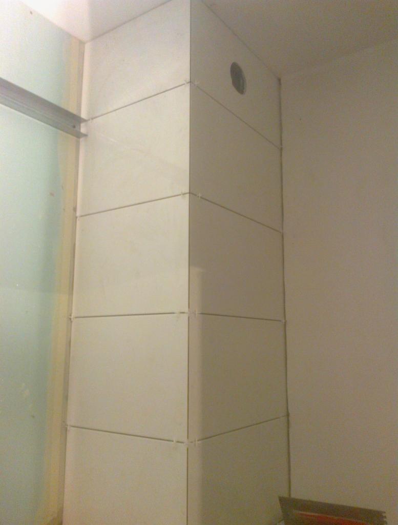 Wykończony narożnik w łazience, obłożony jasnymi, kwadratowymi płytkami ceramicznymi z widocznymi krzyżykami dystansowymi, z otworem wentylacyjnym w górnej części i metalowym profilem po lewej...