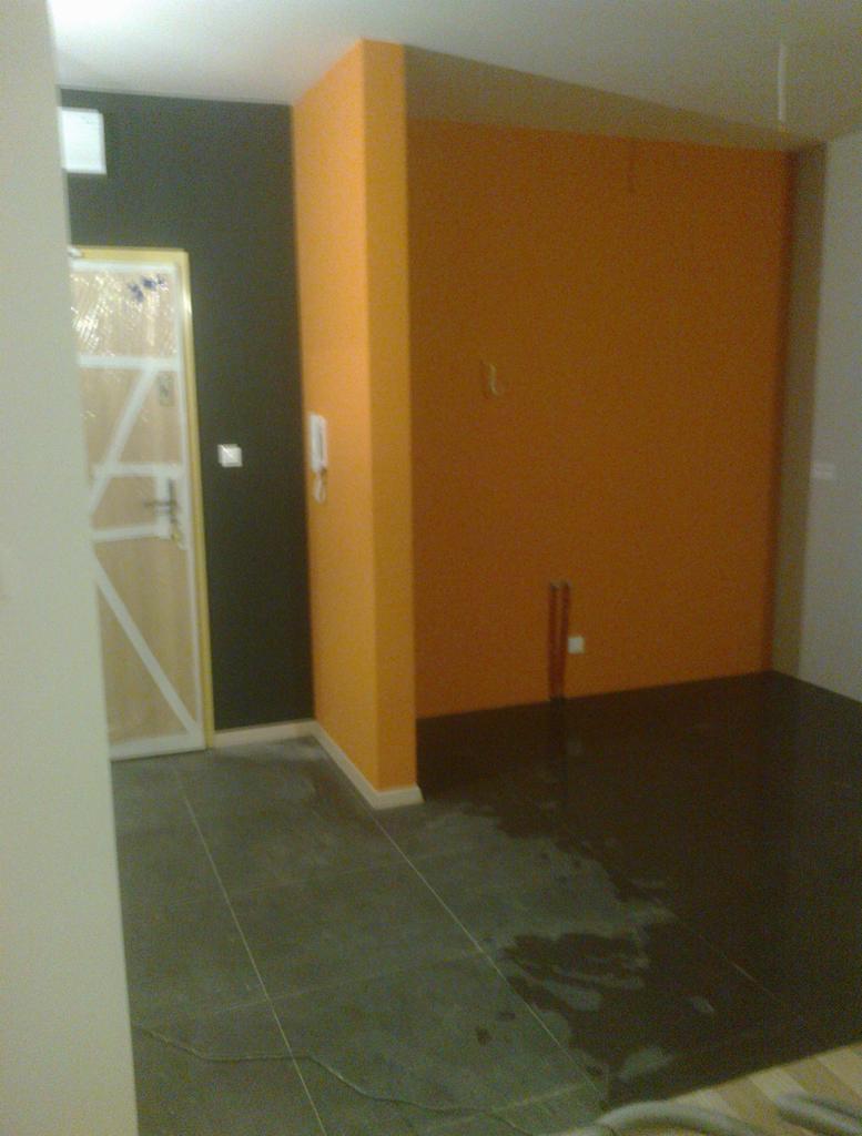 Wnętrze po remoncie z nową, pomarańczową ścianą, ciemną podłogą z płytek i drzwiami zabezpieczonymi folią, widoczne ślady wilgoci na podłodze.