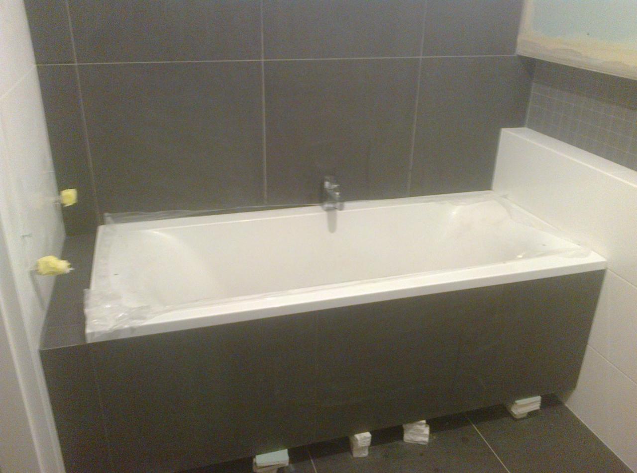 Nowa, biała wanna obudowana szarymi płytkami, zabezpieczona folią ochronną, zamontowana w łazience z szarymi płytkami na ścianach, widoczne kliny pod wanną.