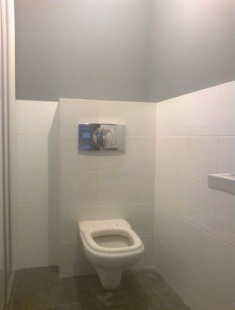 Wisząca muszla klozetowa bez deski na tle białych płytek, z widocznym chromowanym przyciskiem spłukiwania na perforowanej ścianie.