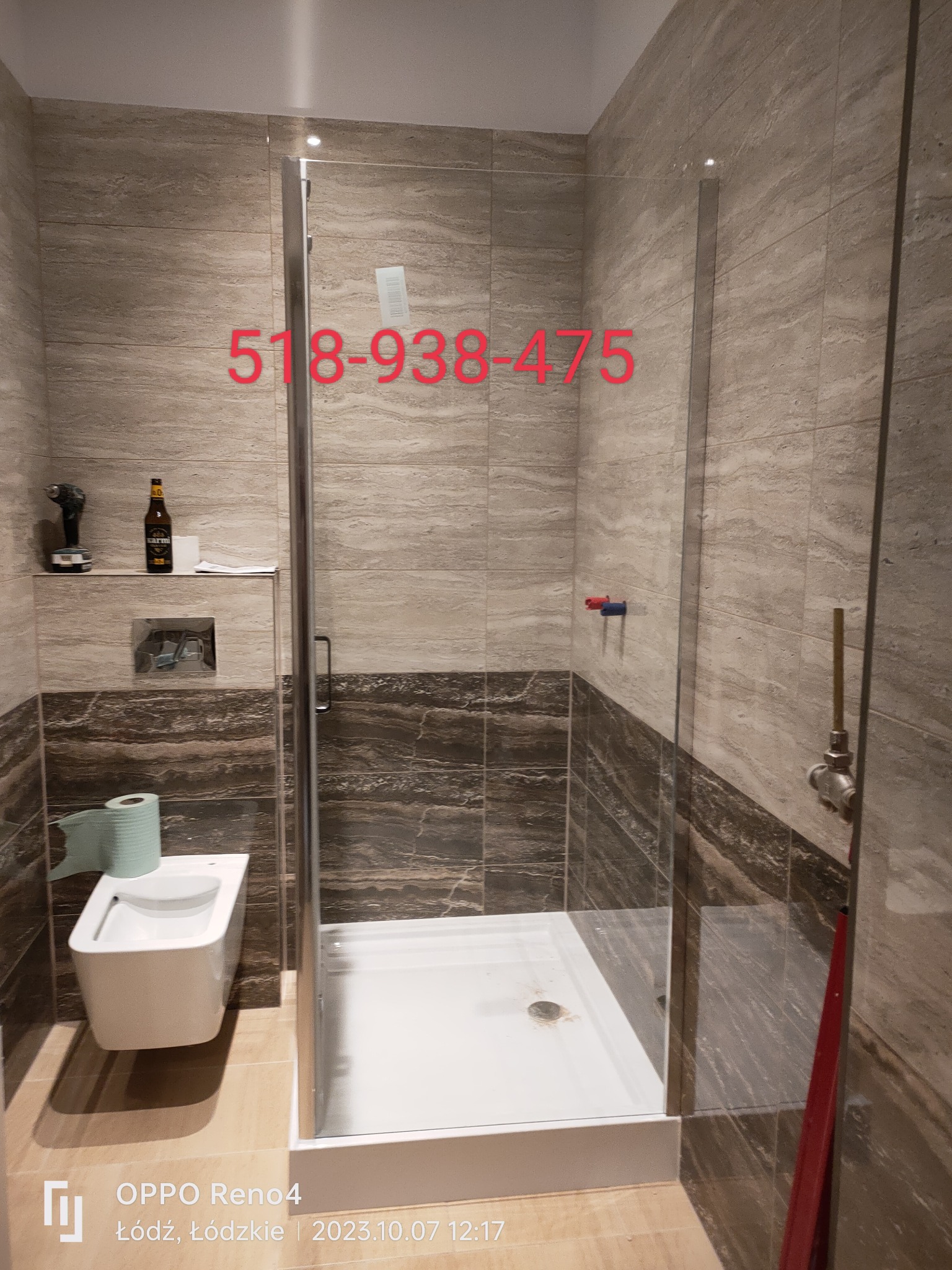 Wykończona łazienka z kabiną prysznicową typu walk-in, wiszącym bidetem, płytkami imitującymi trawertyn oraz półką z wiertarką i butelką piwa. Na szybie kabiny widoczny numer telefonu.