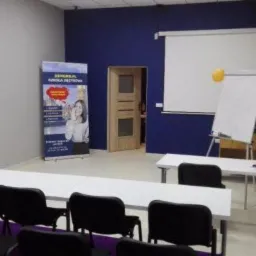 Sala szkoleniowa z białymi stołami, czarnymi krzesłami, tablicą flipchart z żółtym balonem, projektorem i banerem reklamowym szkoły językowej.