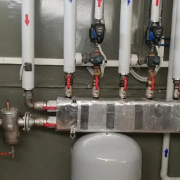 Rozdzielacz ogrzewania podłogowego z pompami obiegowymi, zaworami i izolacją termiczną, widoczne rury z oznaczeniami kierunku przepływu.