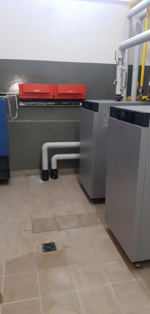 Dwa szare piece gazowe Viessmann z podłączonymi białymi rurami, zainstalowane w pomieszczeniu z kaflami podłogowymi i dwoma czerwonymi panelami sterowania na ścianie.