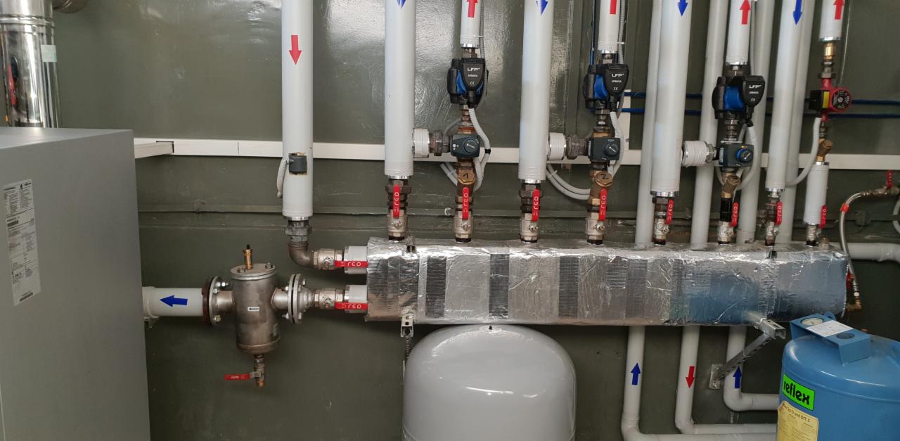 Rozdzielacz ogrzewania podłogowego z pompami obiegowymi, zaworami i izolacją termiczną, widoczne rury z oznaczeniami kierunku przepływu.