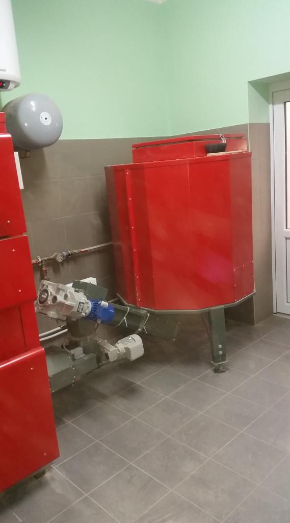 Czerwony, stalowy zasobnik ciepłej wody użytkowej z podłączonymi rurami i silnikiem elektrycznym w pomieszczeniu z szarymi płytkami.