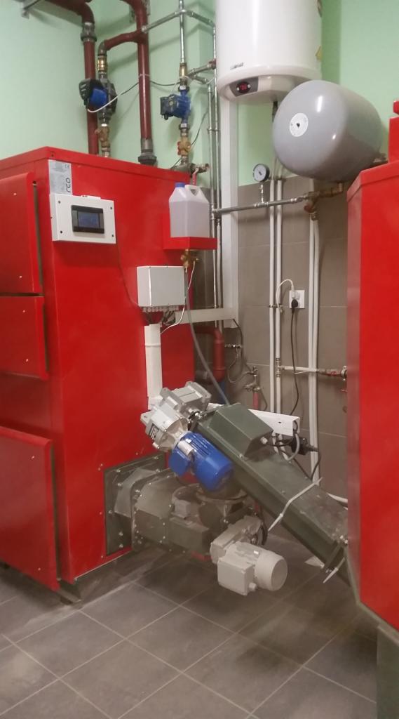 Czerwony piec z podajnikiem ślimakowym, zbiornik wyrównawczy i bojler w kotłowni, widoczne instalacje rurowe i elektryczne.