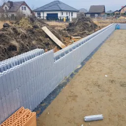 Wznoszenie ścian fundamentowych z szarych bloczków szalunkowych na przygotowanym terenie budowy, widoczne ułożone bloczki z czerwonej cegły oraz w oddali domy jednorodzinne.