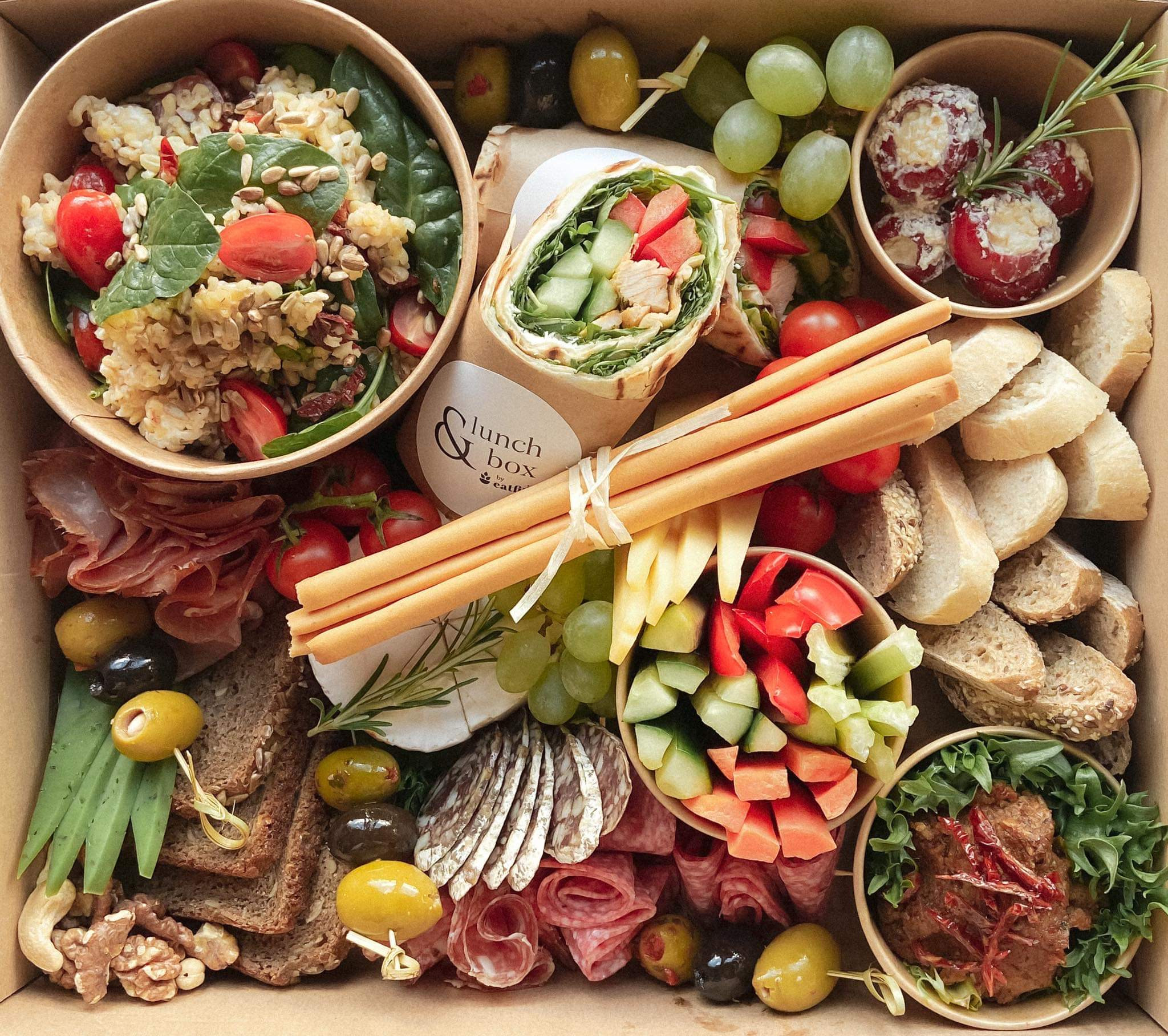 Różnorodna zawartość pudełka z przekąskami: sałatka ze szpinakiem i pomidorami, wrap z kurczakiem, grissini związane wstążką, sery, wędliny, oliwki i winogrona, wszystko starannie ułożone...