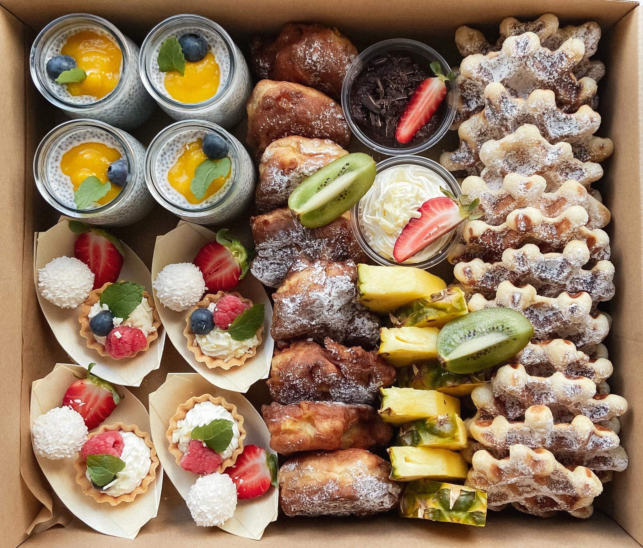 Zestaw różnorodnych słodkich przekąsek w kartonowym pudełku: chia pudding z mango i borówkami, mini tartaletki z kremem i owocami, kokosowe kulki, pączki z cukrem pudrem, pokrojony ananas, kiwi...