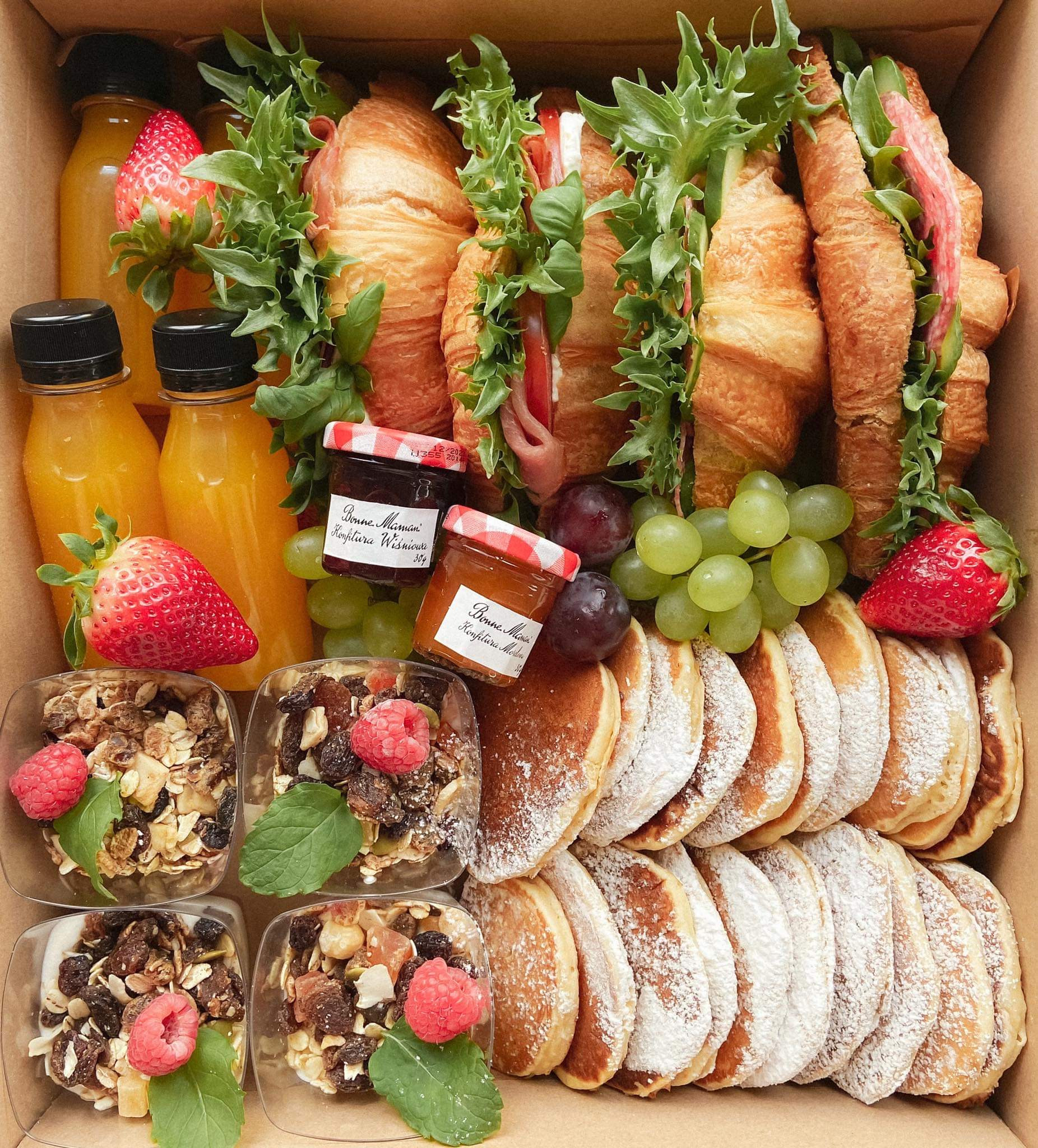 Kartonowe pudełko wypełnione różnorodnym jedzeniem: croissanty z szynką i sałatą, mini buteleczki z sokiem, naleśniki posypane cukrem pudrem, granola z owocami w przezroczystych pojemnikach...