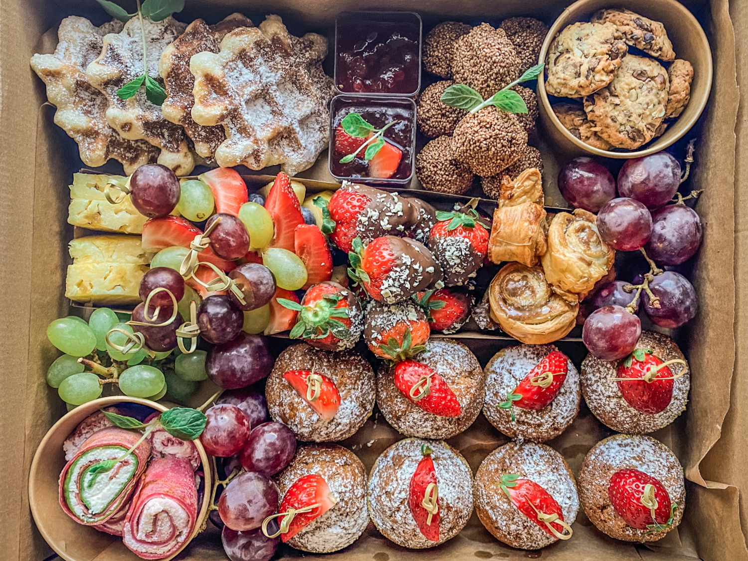 Różnorodna kompozycja słodkich przekąsek w kartonowym pudełku: gofry oprószone cukrem pudrem, kulki czekoladowe, ciastka, winogrona, truskawki (część z czekoladą), roladki z łososiem, ser i małe...