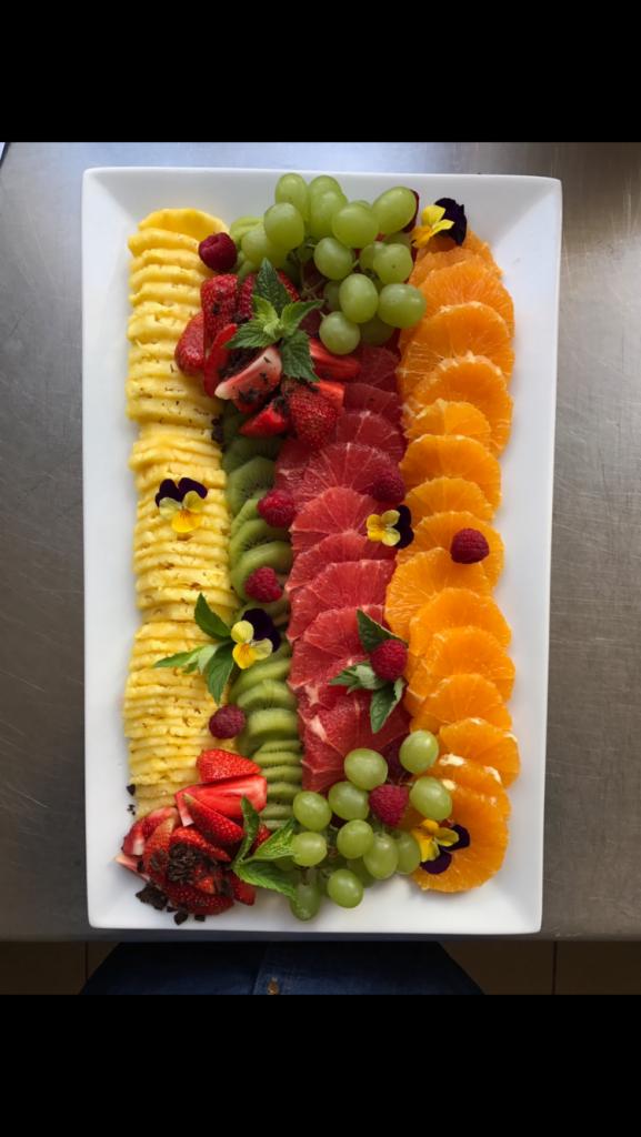 Prostokątna taca z ułożonymi rzędami plastrów ananasa, kiwi, grejpfruta i pomarańczy, udekorowana winogronami, truskawkami, malinami, miętą i jadalnymi bratkami.