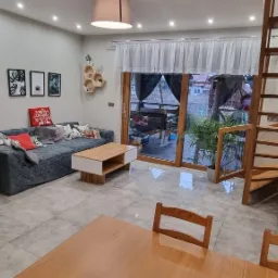 Jasne wnętrze salonu z kominkiem z szarej cegły, narożną sofą z poduszkami, drewnianym stolikiem kawowym, schodami na piętro i wyjściem na taras.