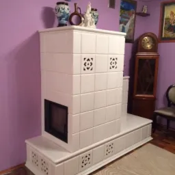 Biały, kaflowy kominek w salonie z fioletowymi ścianami, ozdobiony wazonami i ceramiczną dekoracją na górze, widoczny zegar stojący w tle.