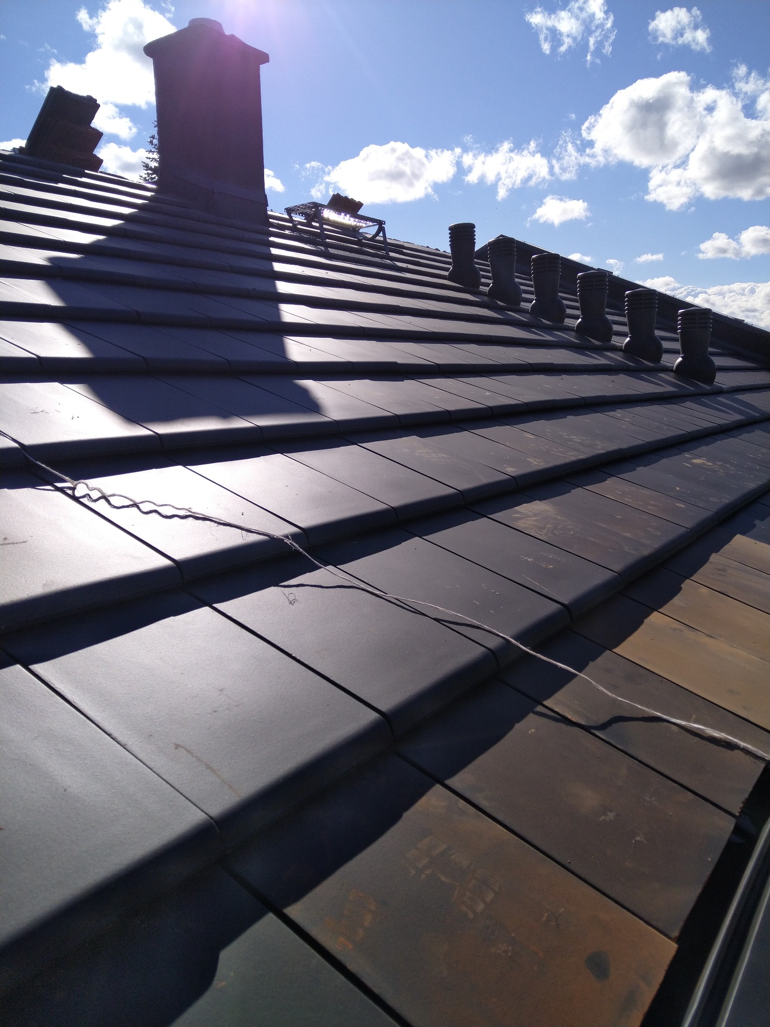 Ciemna dachówka ceramiczna na skośnym dachu w słoneczny dzień. Widoczny komin, wentylacja i fragment instalacji odgromowej. Tło: błękitne niebo z chmurami.