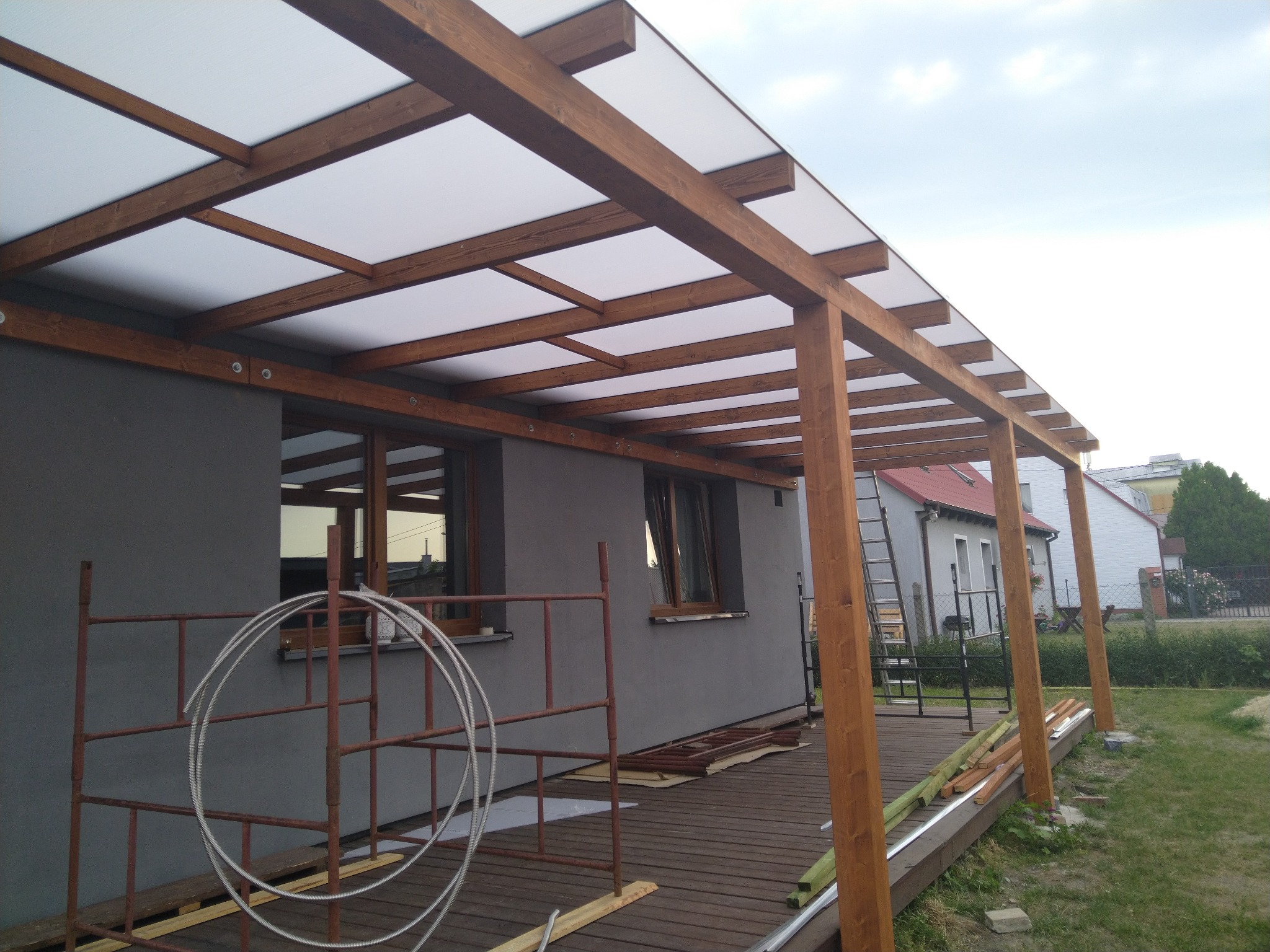 Drewniana konstrukcja z białym zadaszeniem, prawdopodobnie tarasu, w trakcie budowy. Widoczne rusztowania, elementy konstrukcyjne i otoczenie domu.