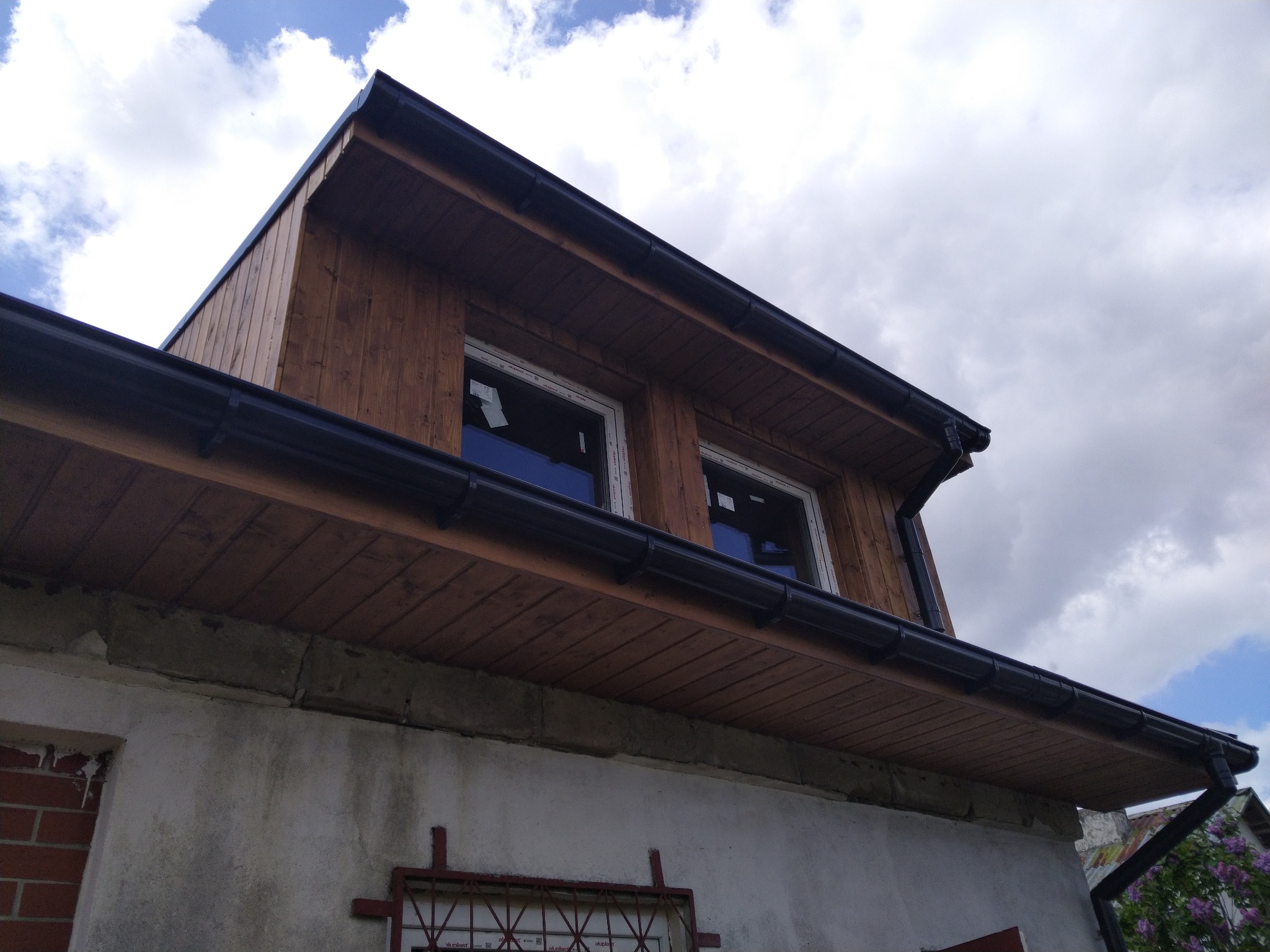 Wykończona lukarna z drewnianą elewacją i czarnymi rynnami, widok z dołu na tle pochmurnego nieba. Detal orynnowania i obróbek blacharskich.