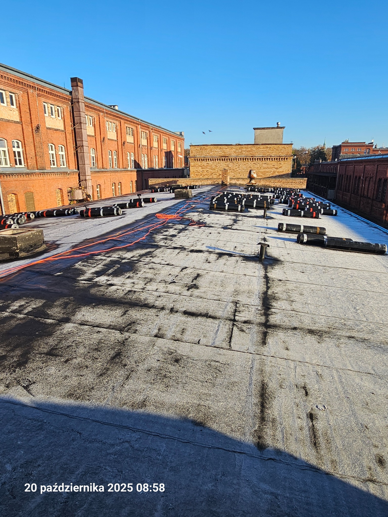 Rozległy, płaski dach budynku w trakcie prac. Widoczne rolki papy, pomarańczowy przewód i elementy wentylacyjne. W tle ceglany budynek i błękitne niebo.