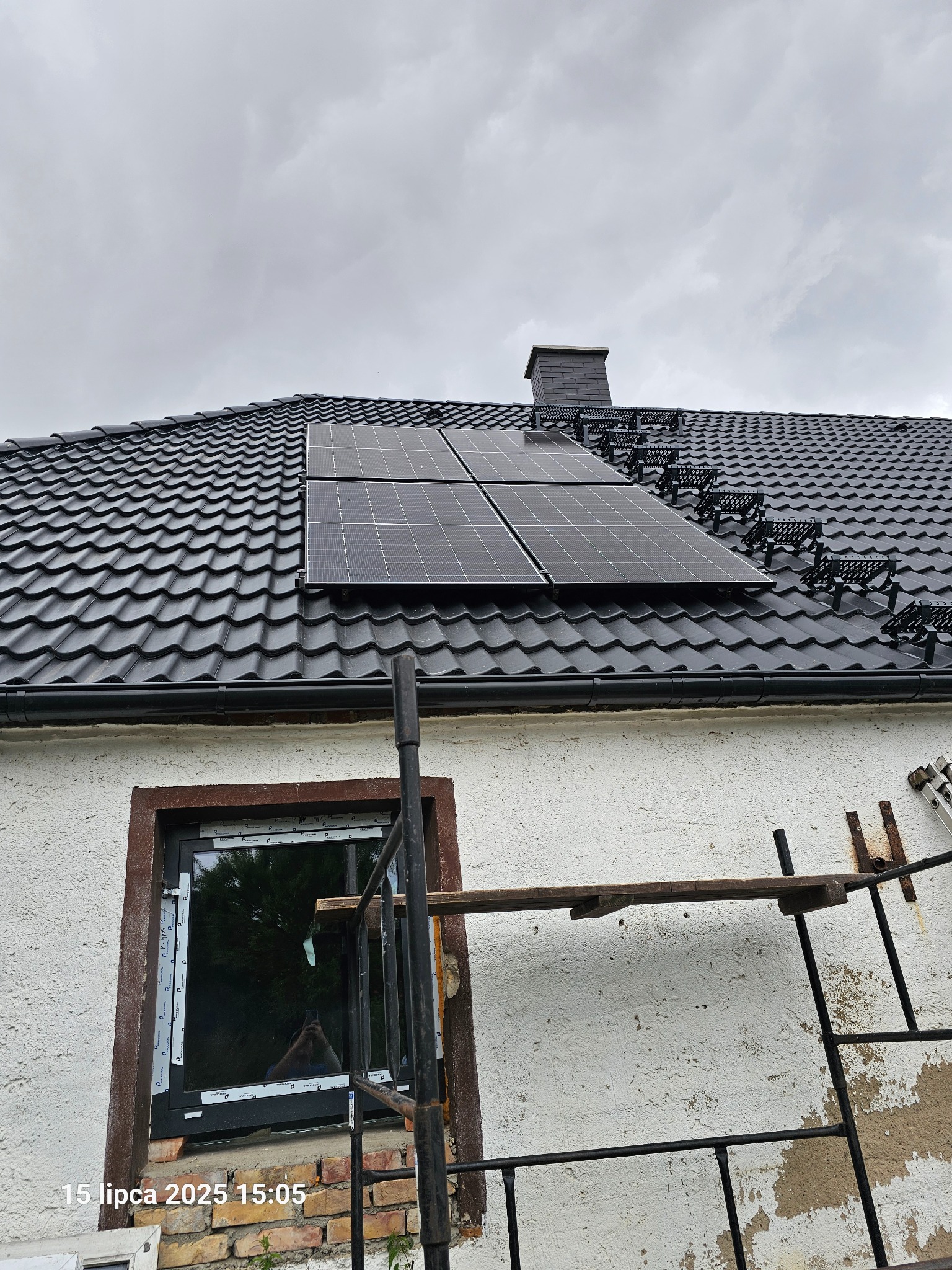 Cztery panele solarne na dachu z czarnej dachówki karpiówki, widoczne zabezpieczenia śniegowe. Fragment ściany z oknem i rusztowaniem. Pochmurne niebo.