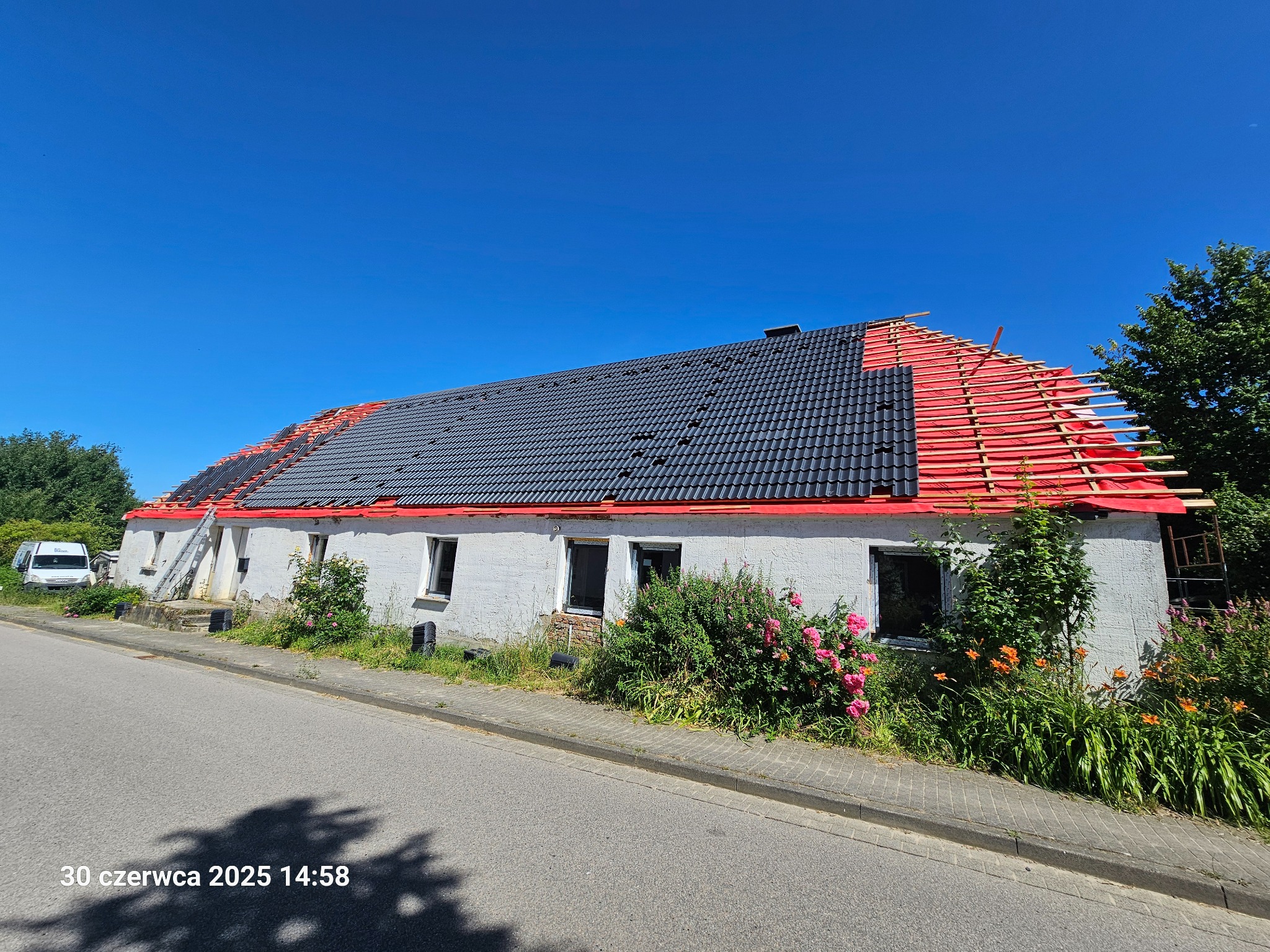 Częściowo pokryty dach czarną dachówką na budynku z białymi ścianami. Widoczne czerwone membrany i drewniana konstrukcja dachu. Część dachu jeszcze bez pokrycia.