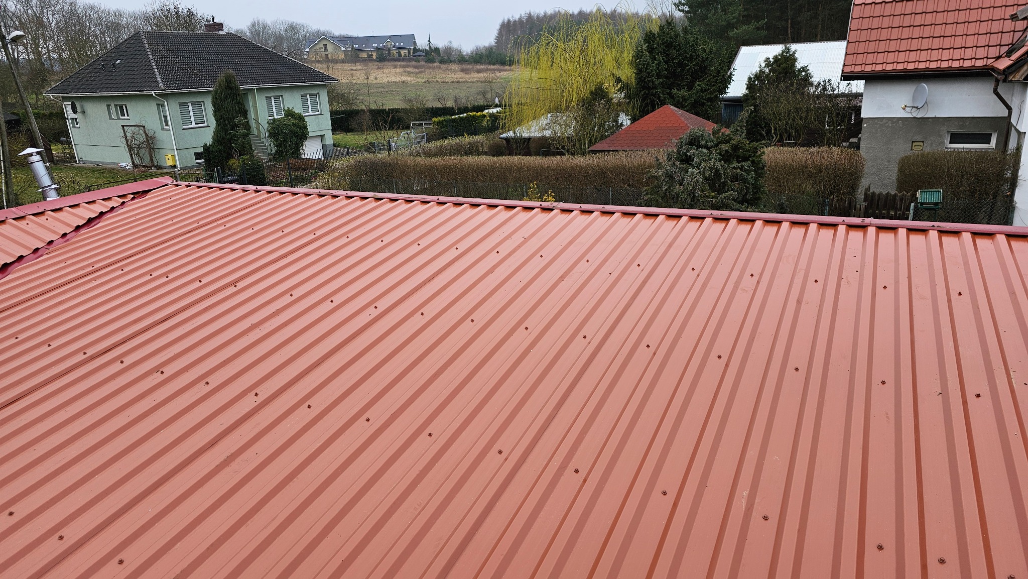 Czerwony dach z blachy trapezowej, widok z góry na tle zabudowy jednorodzinnej i drzew. Nowe pokrycie dachowe z widocznymi śrubami mocującymi.