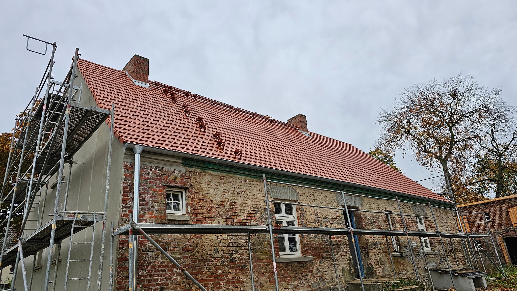 Ceglany budynek z nowym, czerwonym dachem z dachówki, otoczony rusztowaniami. Widoczne kominy i drzewa w tle. Częściowo otynkowana ściana szczytowa.