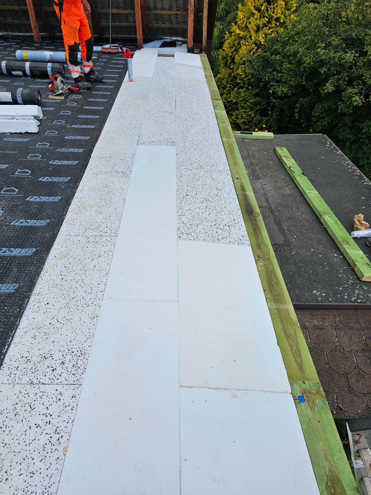 Prace dekarskie na dachu: izolacja z płyt styropianowych układana na membranie. Widoczne narzędzia i dekarz w pomarańczowym kombinezonie. Elementy drewniane wzdłuż krawędzi dachu.