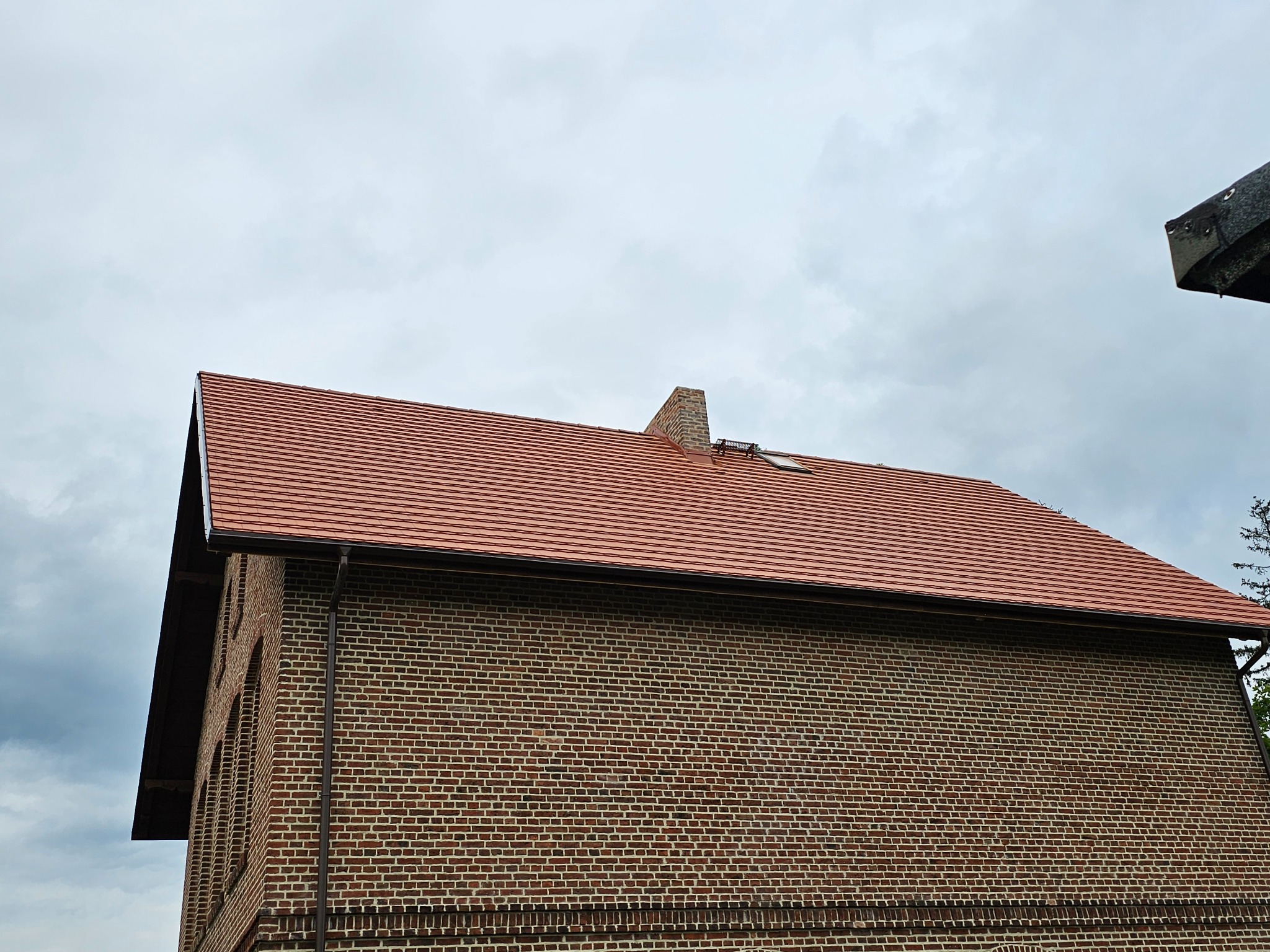 Czerwona dachówka na ceglanym budynku z rynną. Widoczny komin i okno dachowe. Pochmurne niebo w tle. Ujęcie z dołu, podkreślające geometrię dachu.