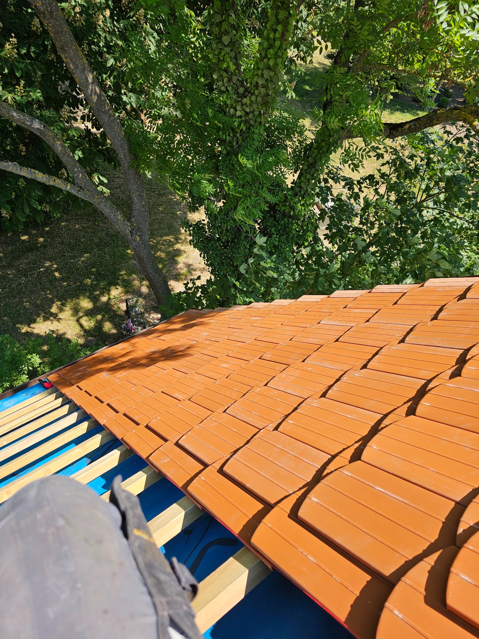 Układanie ceramicznej dachówki w kolorze terakoty na niebieskiej membranie dachowej. Widok z góry, w tle zieleń drzew. Częściowo widoczny element stroju roboczego.