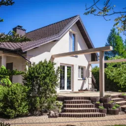 Jasny, dwukondygnacyjny dom z ciemnobrązowym dachem i beżową elewacją, widok z poziomu ogrodu, z tarasem z pergolą i schodami z kostki brukowej. Widoczna zieleń krzewów i drzew.