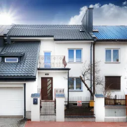 Fragment szeregu domów jednorodzinnych z różnymi typami dachów, od czerwonej dachówki po blachodachówkę w kolorze niebieskim i szarym, białe elewacje, widoczne bramy garażowe i wejścia do domów.