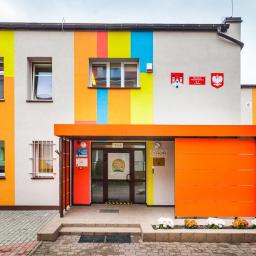 Bautyk - Kolorowa elewacja budynku przedszkola z wejściem osłoniętym pomarańczowym daszkiem i kwiatami wzdłuż chodnika.