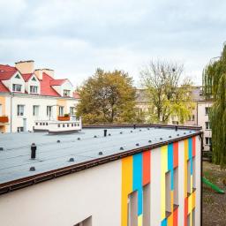 Bautyk - Widok z góry na budynek z płaskim dachem pokrytym papą, fragment elewacji pomalowany w pionowe pasy w kolorach: czerwonym, żółtym i niebieskim, w tle inne budynki i drzewa.