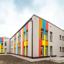 Bautyk - Budynek przedszkola z ocieploną elewacją, pomalowany w pionowe, kolorowe pasy: czerwony, żółty, niebieski i pomarańczowy, z szarą elewacją bazową. Widoczne okna z dekoracjami dziecięcymi.