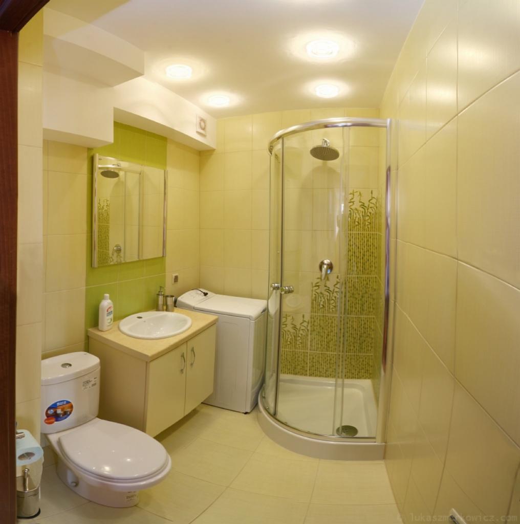 Jasna łazienka w odcieniach żółci z kabiną prysznicową, umywalką z szafką, toaletą i pralką. Widoczne płytki na ścianach i podłodze, okrągłe oświetlenie sufitowe.