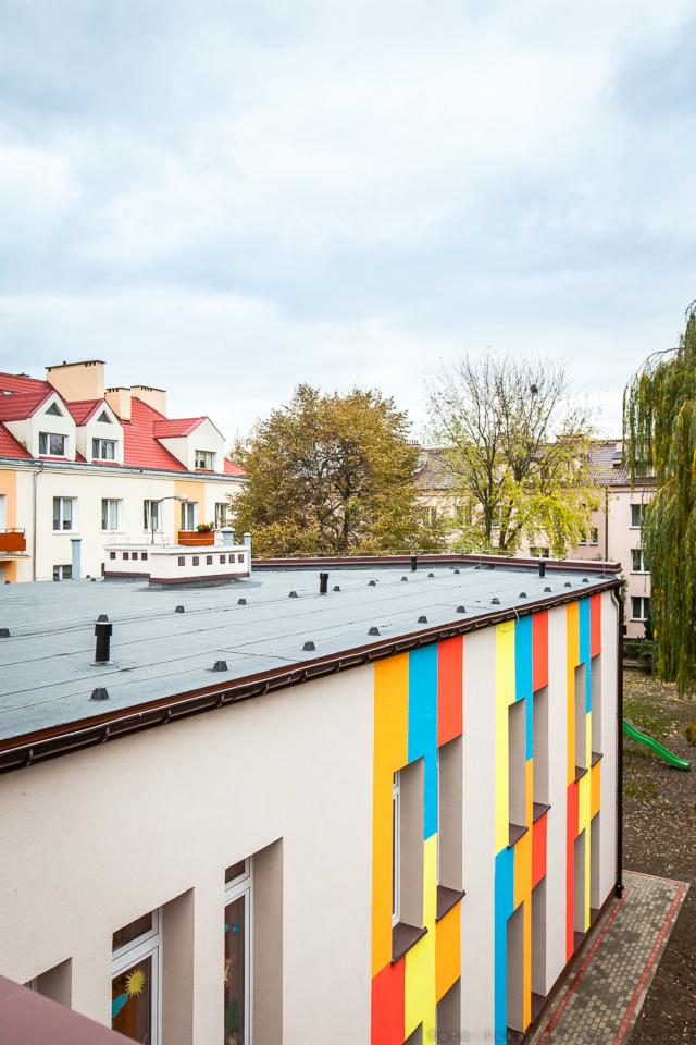 Widok z góry na budynek z płaskim dachem pokrytym papą, fragment elewacji pomalowany w pionowe pasy w kolorach: czerwonym, żółtym i niebieskim, w tle inne budynki i drzewa.