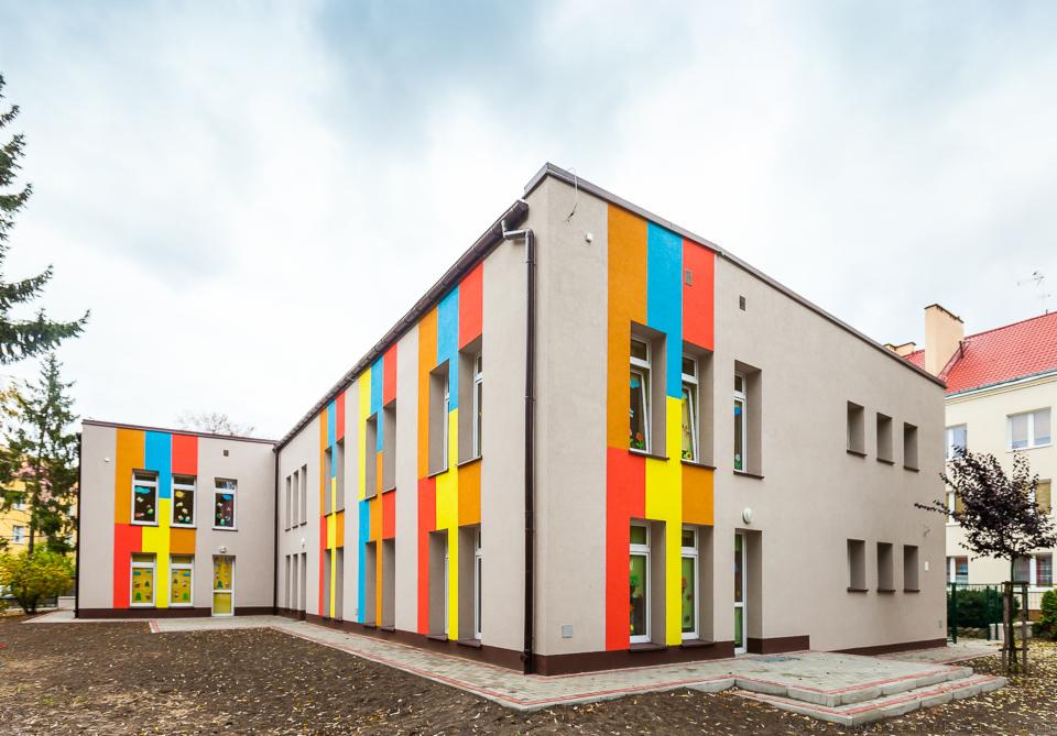 Budynek przedszkola z ocieploną elewacją, pomalowany w pionowe, kolorowe pasy: czerwony, żółty, niebieski i pomarańczowy, z szarą elewacją bazową. Widoczne okna z dekoracjami dziecięcymi.