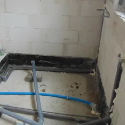 Surowy stan łazienki z widocznymi instalacjami hydraulicznymi w trakcie montażu, ściany z białych bloczków, czarna hydroizolacja przy podłodze, okno z widocznym logo VEKA.