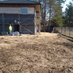 Budowa domu jednorodzinnego w trakcie ocieplania ścian zewnętrznych, widoczne rusztowania, pracownicy w trakcie pracy, teren budowy z ogrodzeniem z siatki.