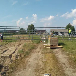 Montaż konstrukcji pod panele fotowoltaiczne na łące, widoczne panele, pracownicy w odblaskowych kamizelkach na drabinach i bez koszulek, wykopana ziemia, błękitne niebo z chmurami.