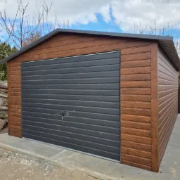 Gotowy garaż blaszany z drewnopodobną elewacją i szarą bramą segmentową, osadzony na betonowej wylewce, w tle zielona roślinność i drewniane palety.