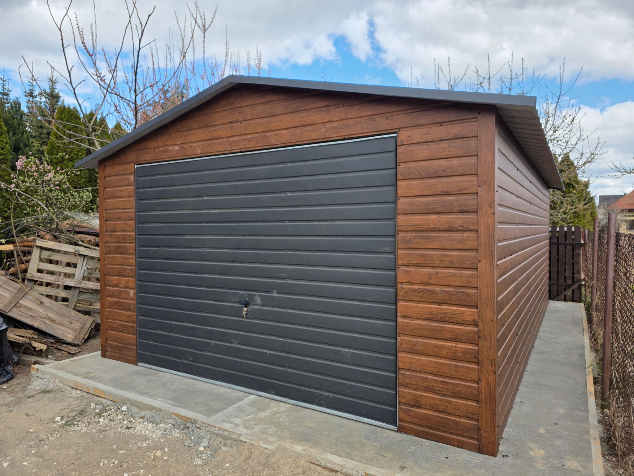 Gotowy garaż blaszany z drewnopodobną elewacją i szarą bramą segmentową, osadzony na betonowej wylewce, w tle zielona roślinność i drewniane palety.